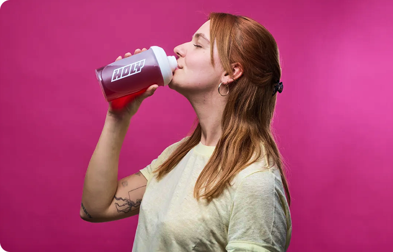 Eine Person trinkt aus einem Shaker vor einem pinken Hintergrund.