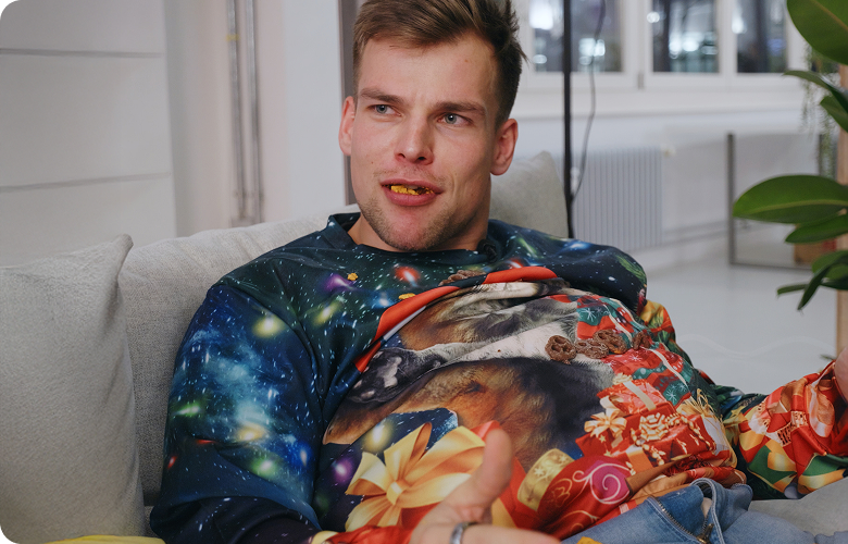 Ein Mann sitzt auf einem Sofa und trägt einen bunten Pullover mit Weltraum- und Wolfsmotiv.