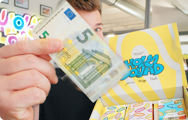 Person hält einen 5-Euro-Schein und zeigt eine gelbe Schachtel mit bunten Probepackungen vor einem Café-Hintergrund.