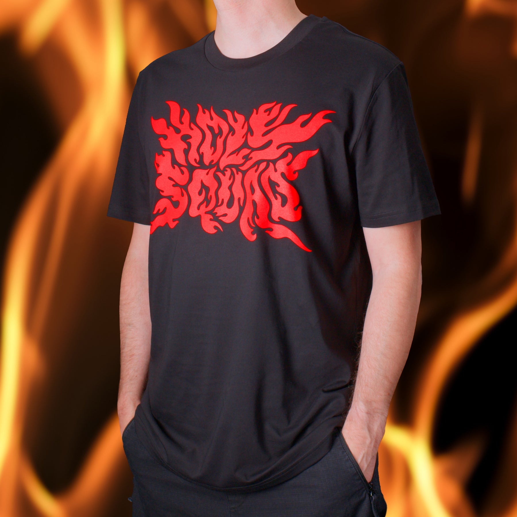 Person in schwarzem T-Shirt mit rotem Flammenmuster, die Hände in den Taschen, vor einem Hintergrund aus Flammen.