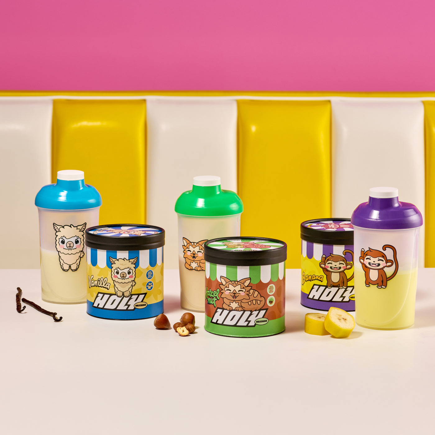 Drei bunte Shaker mit tierischen Motiven und drei Pappdosen mit ähnlichen Motiven auf einem Tisch, umgeben von Vanille, Haselnüssen und Bananen.
