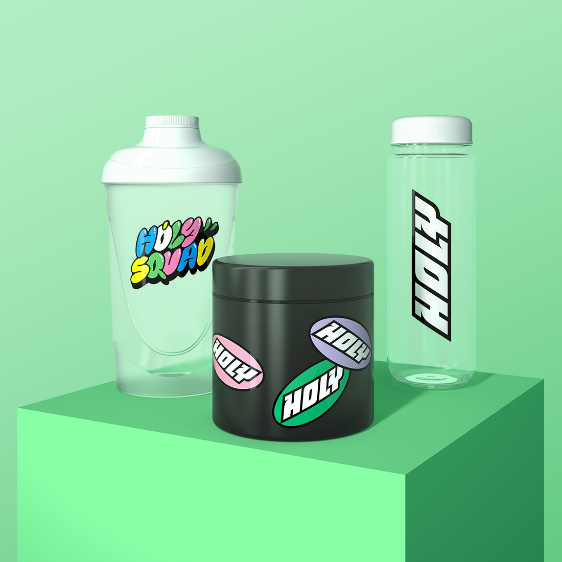 Ein grüner Shaker, eine schwarze Pappdose und ein klarer Shaker mit "Holy"-Design auf einem grünen Podest.