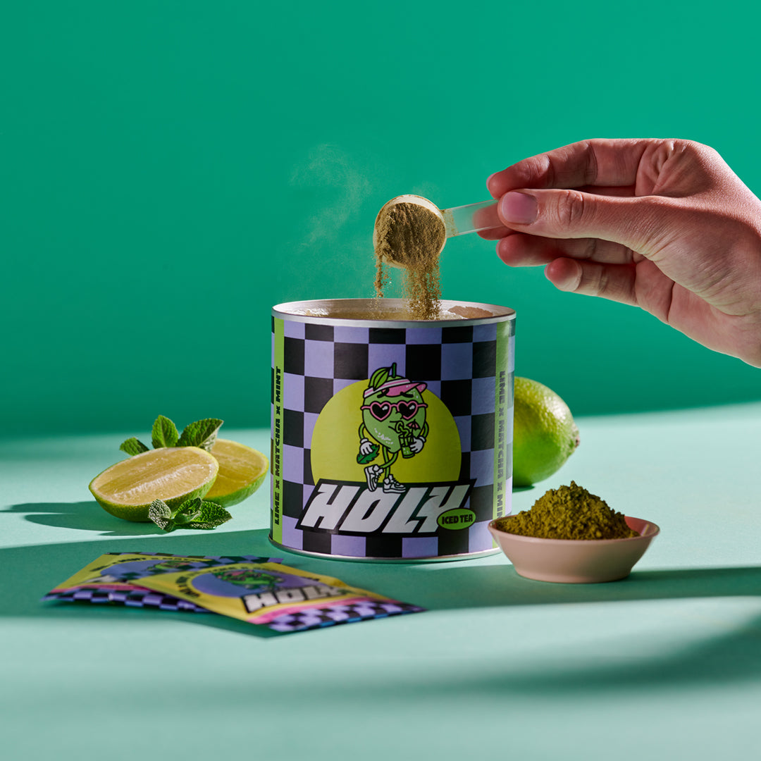 Hand füllt Pulver in eine Pappdose mit limettengrünem Design, daneben Limette, Minze und Probepackungen.