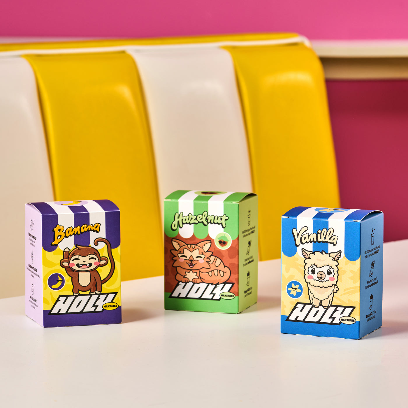 Drei bunte Pappboxen mit Probepackungen der Geschmacksrichtungen Banane, Haselnuss und Vanille auf einem Tisch.
