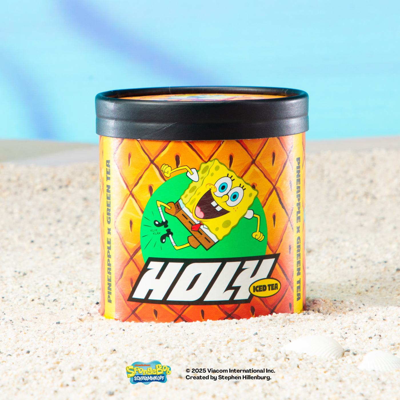 Bunte Pappdose mit Ananas-Design und Zeichentrickfigur, stehend auf Sand vor blauem Hintergrund.