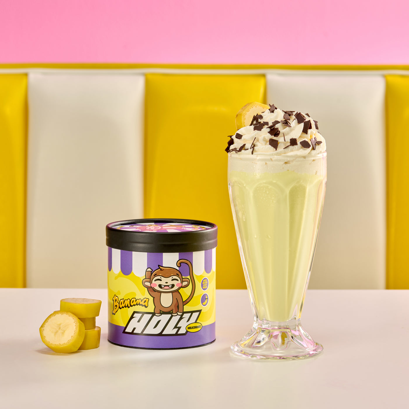 Pappdose mit Affenmotiv und Bananenshake mit Sahne und Schokoladenstreuseln auf einem Tisch vor buntem Hintergrund.