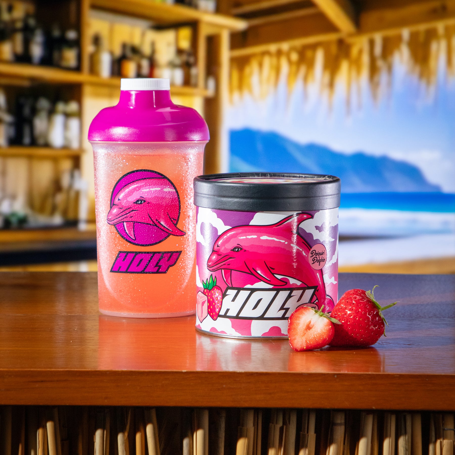 Rosa Shaker und Pappdose mit Delfin-Design und Erdbeeren auf einem Tisch vor tropischem Hintergrund.