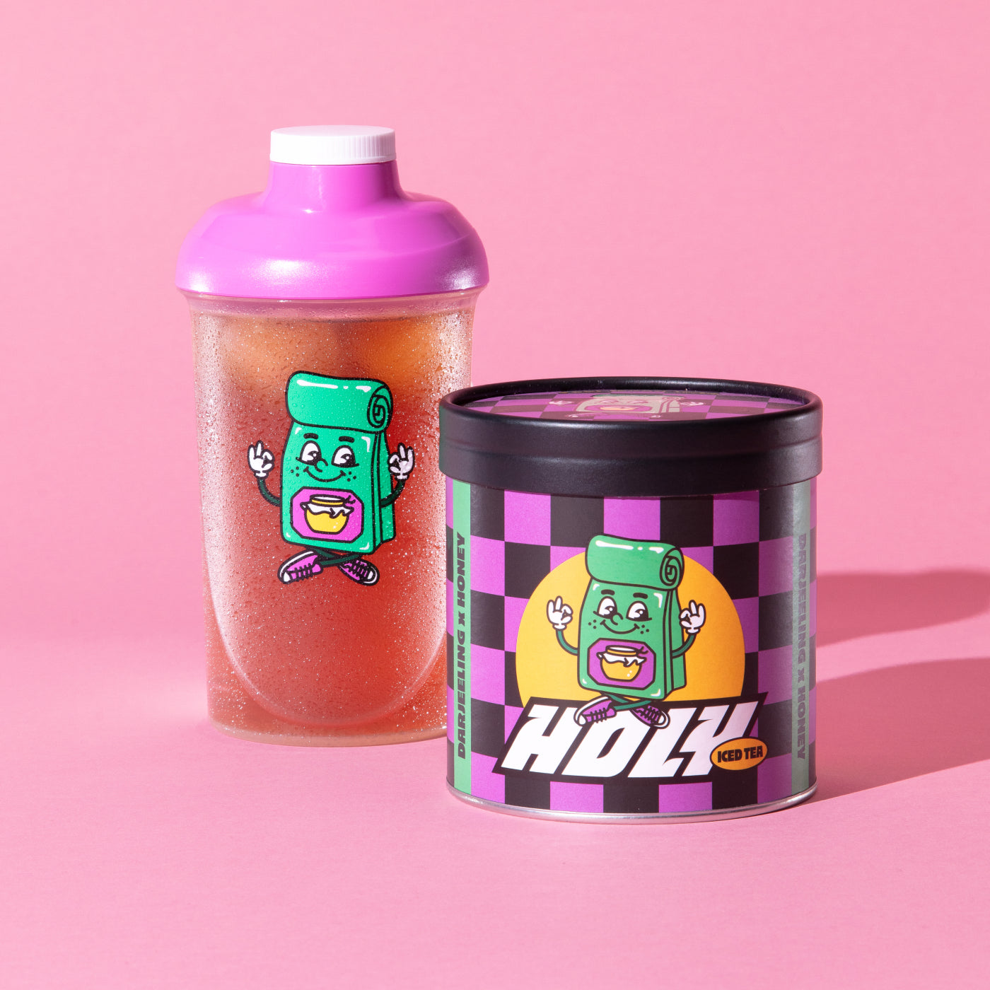 Ein pinker Shaker und eine karierte Pappdose mit buntem Cartoon-Aufdruck vor rosa Hintergrund.