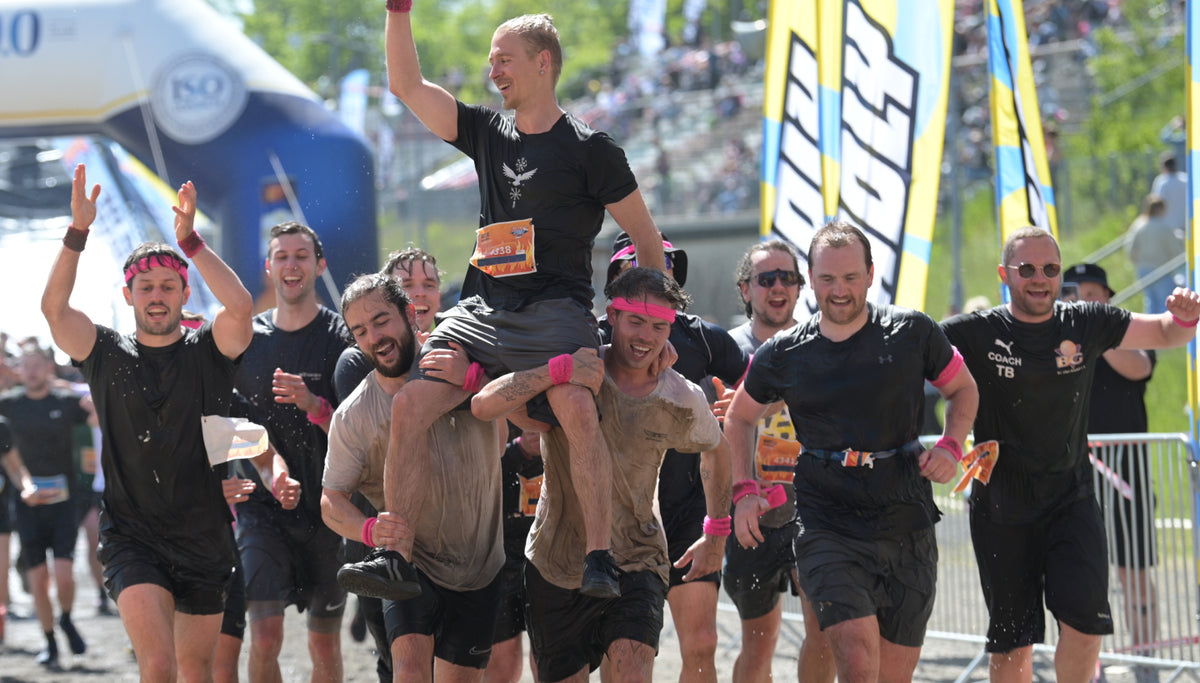 Gruppe lachender Männer in Sportkleidung beim Laufwettbewerb, einer wird auf Schultern getragen, umgeben von bunten Fahnen.