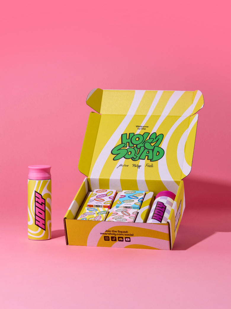 Gelbe Pappbox mit mehreren Probepackungen und einem gelben Shaker auf rosa Hintergrund.