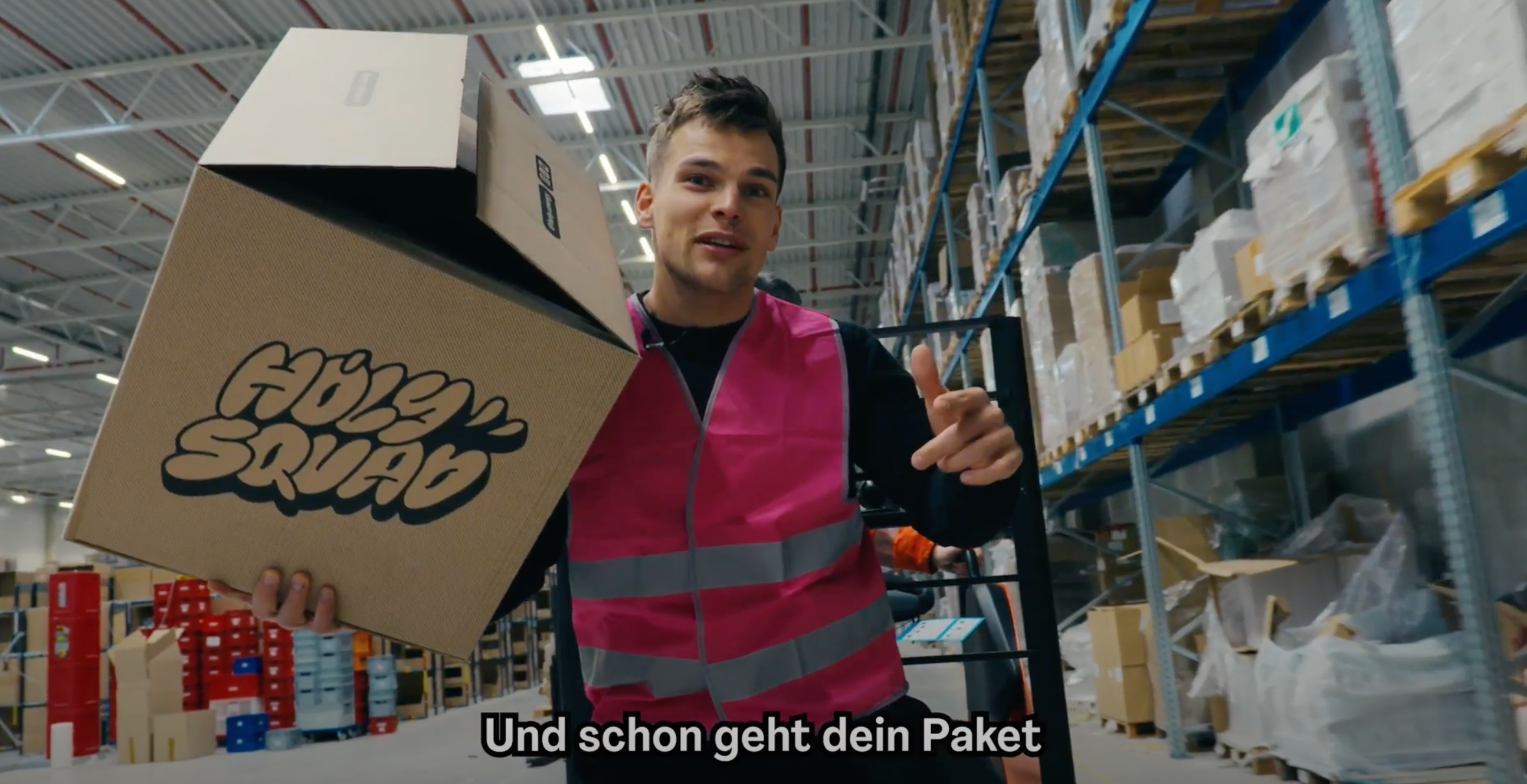 Ein Mann in einer Lagerhalle trägt eine große Pappbox mit der Aufschrift "Holy Squad" und eine pinke Weste.
