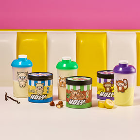 Drei bunte Shaker mit tierischen Motiven und drei Pappdosen mit ähnlichen Motiven auf einem Tisch, umgeben von Vanille, Haselnüssen und Bananen.