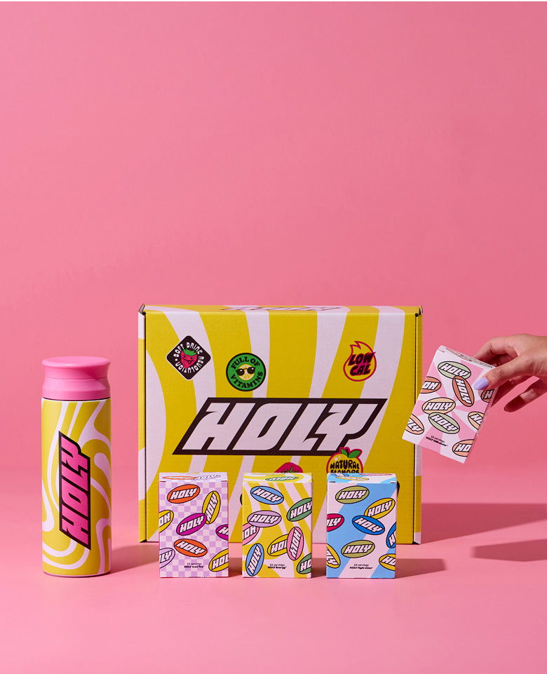 Gelb-pinke Pappdose, Schachtel mit HOLY-Logo, drei Schachteln und eine Hand mit einem Sachet vor pinkem Hintergrund.